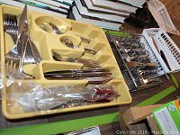 Flatware tray filled with forks, spoons, knives, and steak knives along with other small utensils shown in a corner with books and baskets nearby.