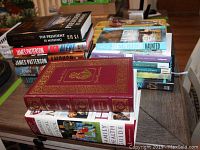 Multiple hardcover books stacked on a table, including titles by James Patterson, Bill Clinton, John Grisham, and Nancy Kennedy.