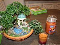Photo of Thomas Kinkade flower garden decorative piece with greenery, gazebo, water feature, and two orange candles on a table.