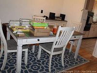 Full view of rectangular dining table with six drawers, distressed white paint, brown wooden top, and matching chairs on patterned blue and white rug.