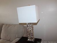 Two matching Ashley table lamps with square white shades and mosaic patterned bases on a dark surface near a gray couch, one lamp shown fully in one image.