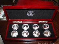 Open red wooden box displaying eight silver coins in individual protective cases, each with different designs.
