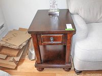 Front view of dark brown wooden side table next to a sofa, showing single drawer and lower shelf