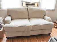 Front view of light beige loveseat with two seat and two back cushions, positioned on a hardwood floor below windows with a window unit air conditioner.