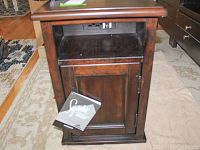 Front angled view of dark wood Ashley Furniture end table showing the open shelf with power outlets and USB, a lower cabinet door, and attached Lynn Signature Design booklet