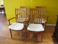 Four teak wooden captain's chairs grouped corner with yellow wall background, showing vertical slat backs and cushioned fabric seats.