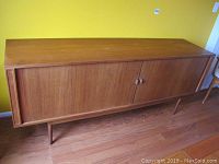 Front left perspective of teak buffet sideboard with sliding doors on wooden legs against a yellow wall