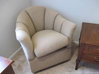 Beige semi round upholstered glider chair positioned near a corner with wooden side tables around it, showing rounded padded backrest, armrests, and a skirted base.