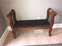 Front view of light brown wooden bench with dark upholstered seat and swirl pattern in a tiled room.