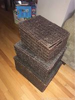 Photo showing three stacked rectangular dark brown wicker baskets in used condition on a hardwood floor near a sofa and white wall.