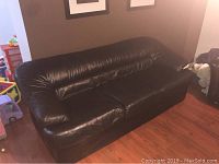 Full view of a black faux leather couch with two seat cushions and padded armrests, placed on wooden floor.