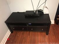 Front view of black wood TV stand showing open middle shelf and two closed drawers with silver knobs, placed on hardwood floor.