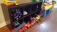 Dark wood cubby shelf holding toy fire trucks, cars, and a yellow construction truck on floor in front.