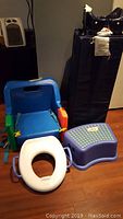 Photo showing the blue highchair seat, white toilet training seat, and purple step stool arranged on wooden floor with playpen in background.