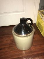 Large ceramic jug style growler placed on wooden floor in front of a door with a pale yellow crate to the side.
