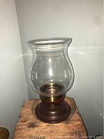 Photo of a single hurricane lamp with a clear glass chimney, brass holder, and dark wooden round base, placed against a white background.