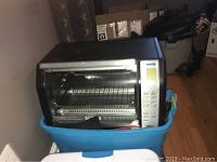 Black convection oven sitting on a blue plastic container, showing the front with door and rotisserie inside.