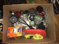 Top view of cardboard box containing assorted kitchenware including travel mugs, glasses, coffee filters, and a wire whisk