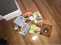 Photo showing assortment of 9 cookbooks on wooden floor and on table with visible titles and cover images.