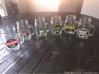 Six clear glass mason jar mugs with Christmas phrases arranged on a dark wooden table.