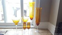 Front view of amber glass stemware and vases grouped on floor in front of patio door showing variety of glass shapes and heights