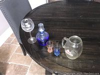 Coors Light mug, blue decanter, etched wine glass, and colored shot glasses arranged on a round wooden table.
