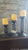 Front view of a set of three metal spiral-style candle holders with yellow pillar candles of varying sizes against a stone wall background.