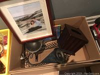 Box containing various house decor items like a framed photograph, wooden Kleenex holder, black metal basket, and some metal decorative pieces