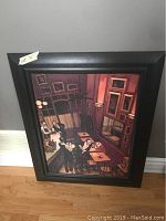 Framed art print showing a bar scene with people inside a room, leaning against a wall on a hardwood floor.