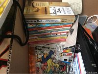Box with stacked books including 'Heads in Beds' and other assorted books and magazines, a visible cardboard tag and a small bottle present