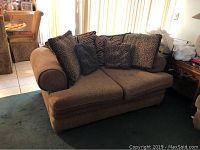 Full view of brown upholstered loveseat with five animal print decorative pillows, showing overall wear and stains.