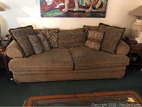 Full view of the large three-seater brown upholstered couch with six animal print pillows on top, placed on a green carpet near wooden furniture.