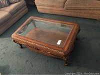 Full view of wooden coffee table with glass top showing carved decorative edges and legs, sitting on carpeted floor in front of couches.