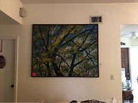 First photo shows one large oil painting on canvas hanging on a white wall above a dark surface. The painting depicts tree branches and foliage in green, yellow, and blue colors in an abstract style.