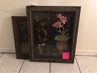 Two framed 3D floral metallic wall pictures leaning against a wall on a tiled floor. The front picture features a brown ornate frame with a large vase holding pink flowers and smaller flowers in panels.