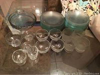 Wide shot showing 13 clear glass dessert serving cups, 29 round glass plates stacked, and 2 large serving trays