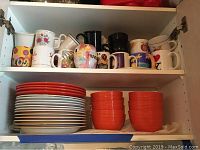 Stack of plates and assortment of colorful mugs on two cabinet shelves.