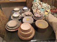 Variety of decorated plates and bowls arranged on a table showing multiple stacks and single plates, including Mikasa floral patterns and other decorated pieces.