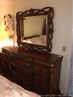 Wooden dresser with multiple drawers beneath a large decorative wooden framed mirror, showing overall condition and wear