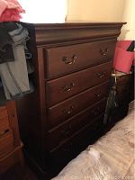 View of five drawer wooden dresser in dark brown finish with brass handles, showing signs of wear.