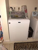 Full view of white top load washing machine with control panel visible, and some surrounding laundry area.