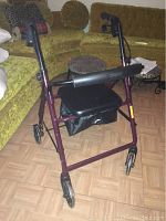 Photo of maroon four-wheel rollator walker with padded armrests, seat and back support, positioned indoors on wooden flooring near green couch
