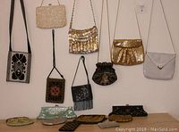 Wide shot showing thirteen assorted vintage evening bags and purses hanging on wall and lying below in one group. Bags include beaded, metal mesh, fabric, and sequin styles in various colors and designs.
