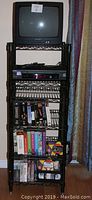 Full view of black 6-shelf adjustable storage rack with small CRT TV on top, Sony VHS player on second shelf, and multiple VHS tapes on lower shelves.
