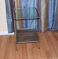 Side view of silver gray metal side table showing glass top and slatted metal lower shelf, standing on wooden floor with striped curtains in background.
