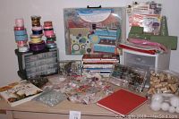 Craft supplies including watercolor art books, ribbons stacked on an organizer, plastic storage bins, assorted beads, jewelry supplies, corks, foam balls, and gift bags visible on a table.