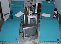 Full front view of two turquoise Sharper Image lounge chairs with armrests and white spa pillows. Positioned between the chairs are a small CRT TV, a metallic mini fridge, a brass ice bucket, a blue water bottle, a boxed wine opener, and a decorative item.