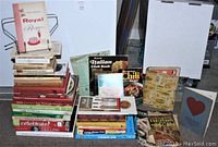View of the entire assortment showing stacked contemporary and vintage cookbooks with a metal cookbook holder behind them.
