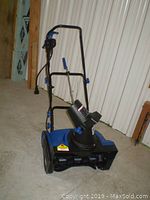 Front full view of the blue and black Snow Joe electric snowblower standing on concrete floor with handle upright and chute facing forward.