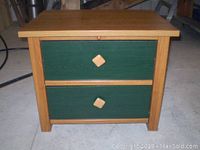 Front view of the side table showing two green painted drawers with wooden diamond-shaped knobs and natural wood frame.
